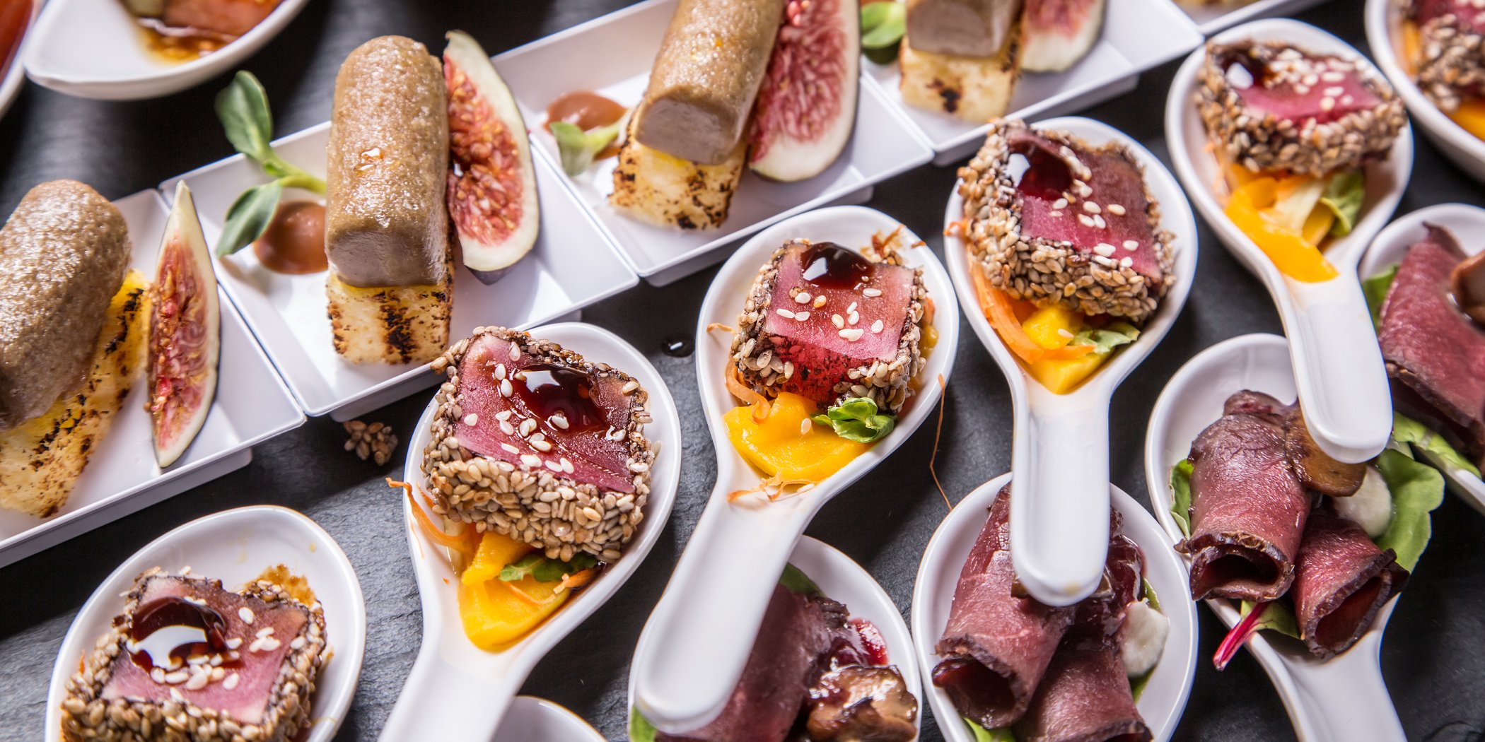 Large presentation spoons containing a selection of roast beef appetisers cooked rare and accompanied with sesame seeds and extravagant fruits