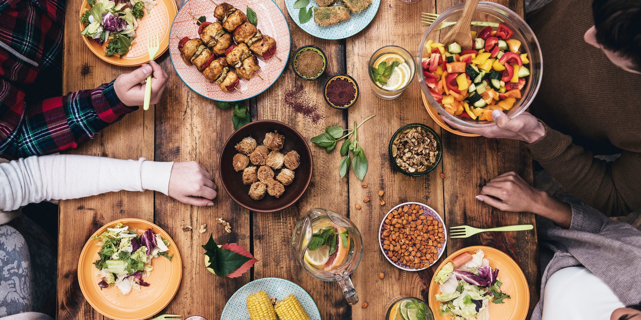 Rustic table surrounded by group of friends socialising and eating tasty homemade food including salad, chicken skewers and sweetcorn
