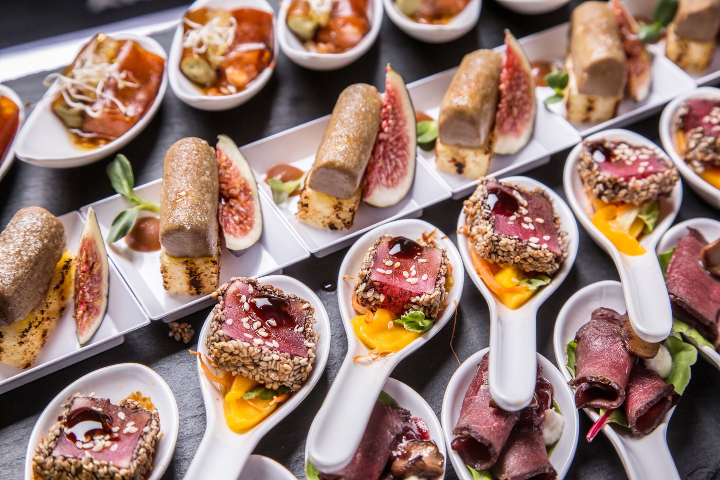 Large presentation spoons containing a selection of roast beef appetisers cooked rare and accompanied with sesame seeds and extravagant fruits