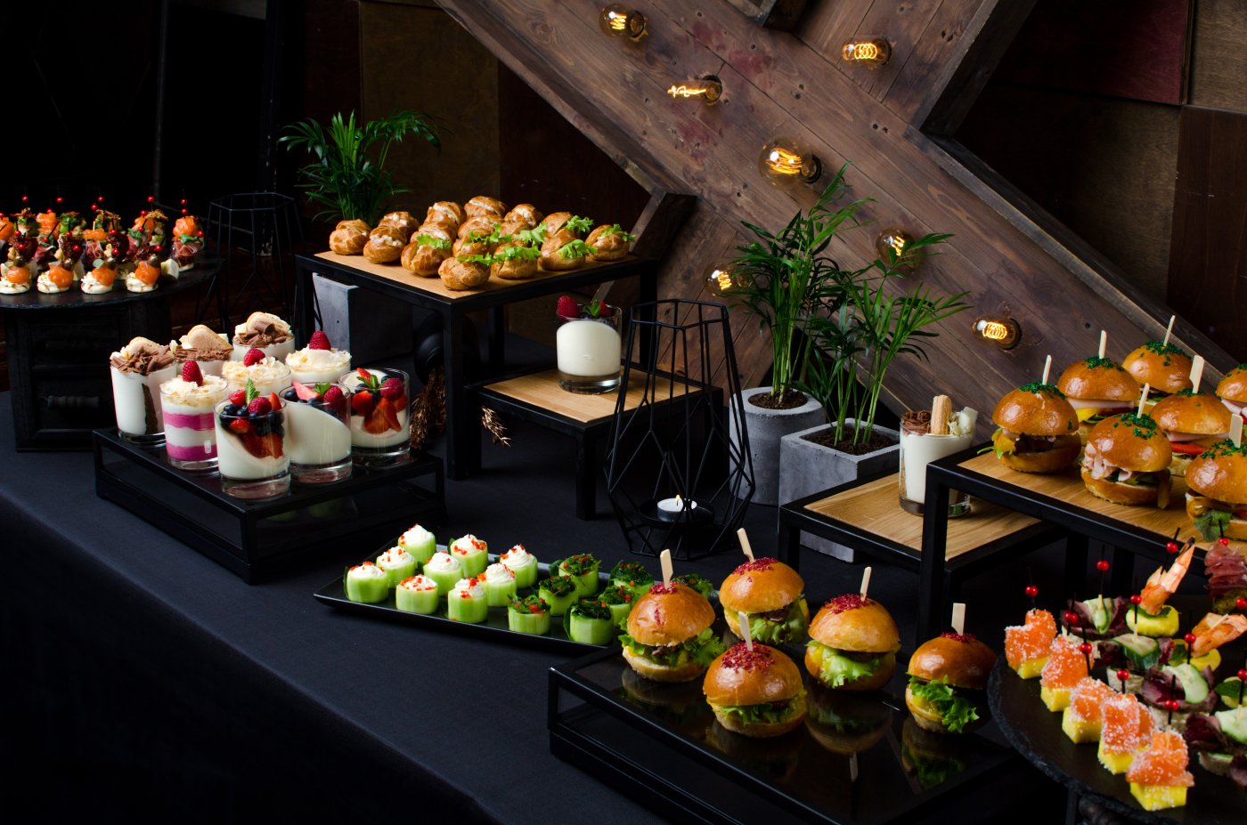 A dark backdrop with a selection of glossy black plates containing various appetisers including mini burgers, deserts and salads