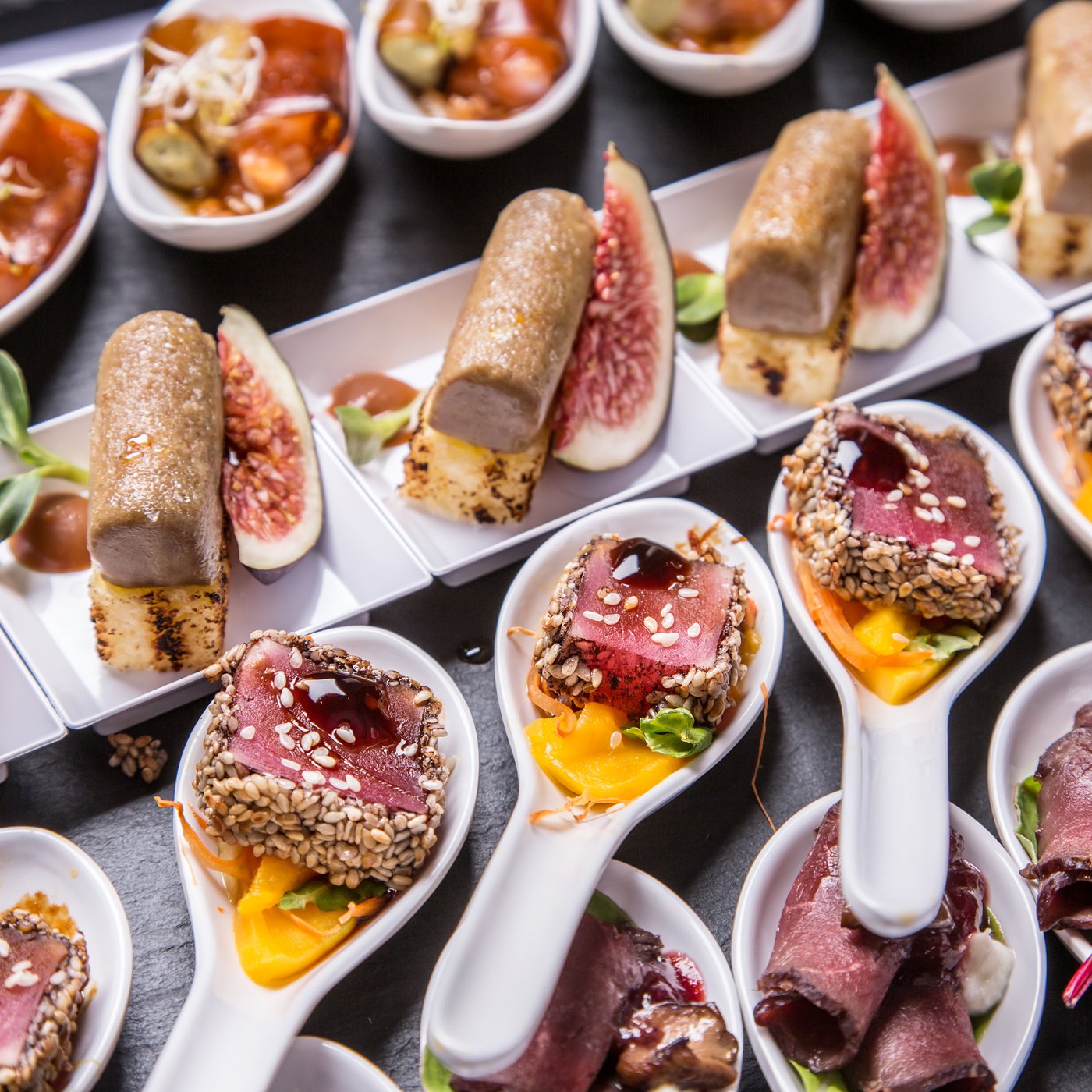 Large presentation spoons containing a selection of roast beef appetisers cooked rare and accompanied with sesame seeds and extravagant fruits