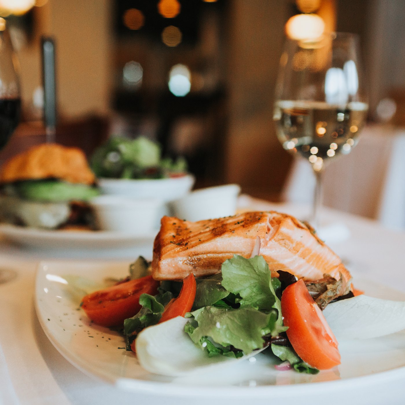 Salmon on a bed of tomato and rocket served on a white dish with a glass of wine in the background